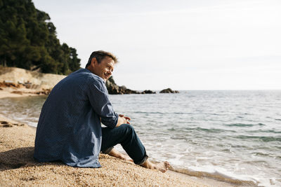 Smiling man sitting on beach while looking away against sea at beach