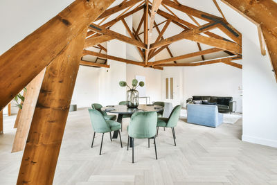 Interior of modern spacious open plan living room with creative wood beams on ceiling furnished with comfortable sofa and armchairs and decorated with carpet and potted plants near dining table