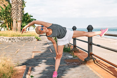 Young beautiful sportive woman training over seaside promenade, stretching legs before jogging. 