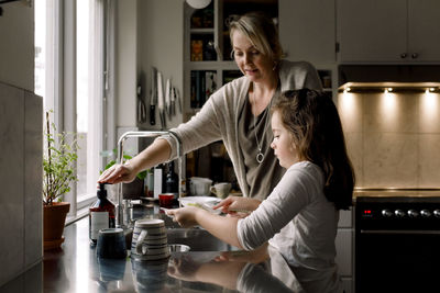 Daughter washing dishes while standing by mother in kitchen at home