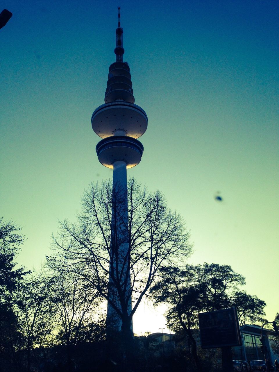 low angle view, tower, building exterior, architecture, tall - high, built structure, clear sky, blue, tree, communications tower, street light, sky, travel destinations, spire, bare tree, fernsehturm, sphere, silhouette, city, tall