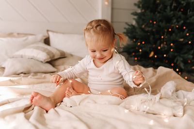 Happy cheerful baby little girl sitting smiling on a cozy bed at christmas holidays at home