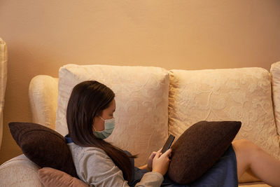 Girl wearing mask using smart phone while lying down on sofa at home