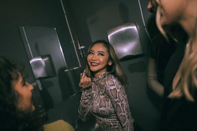 Smiling young woman applying lipstick while looking at friends in nightclub bathroom