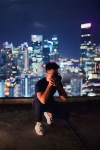 Full length of man photographing on building terrace against illuminated cityscape at night