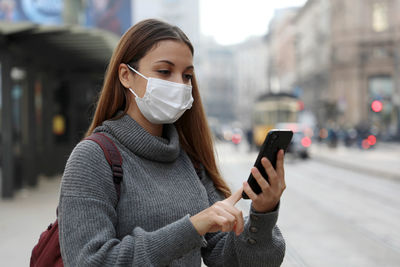 Young woman wearing mask using phone while standing outdoors