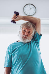 Bearded aged man in blue t shirt breathing and looking at camera while lifting dumbbell over head during fitness workout at home