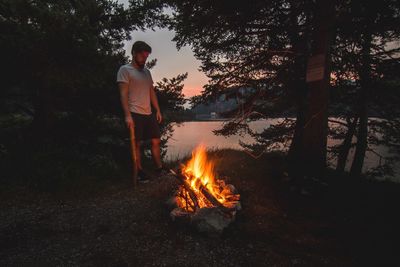 Young man standing by bonfire in forest at dusk