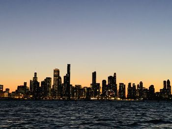 Sea by modern buildings against clear sky during sunset