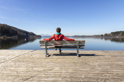 Rear view of man sitting on pier over lake against sky