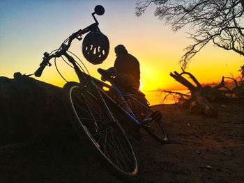 Silhouette man sitting by bicycle against orange sky during sunset