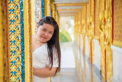 A smiling asian woman in a white dress hides behind a pillar in buddhist temple