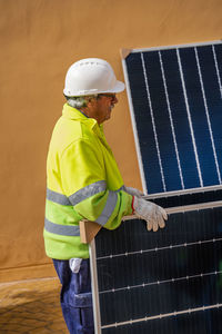 Side view of mature experienced male technician in uniform and helmet standing with solar panel near yellow building while working on installation of renewable energy system