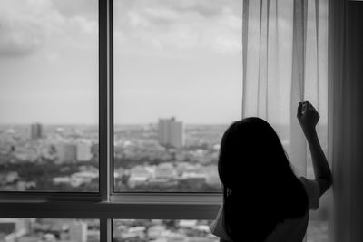 Rear view of woman holding curtain and looking out of glass window. depressed woman from lock down.