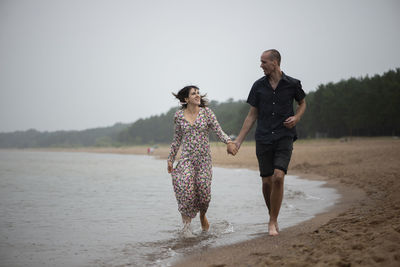 Young happy couple holding hands running along the sea shore ar rainy summer day