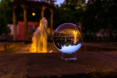 Reflection of illuminated crystal ball on glass against trees at night