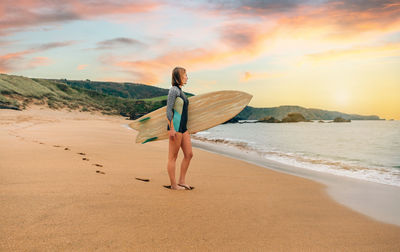 Surfer woman with wetsuit carrying surfboard looking to the sea at the beach