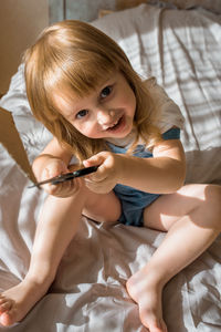 High angle portrait of girl playing with scissors while sitting on bed