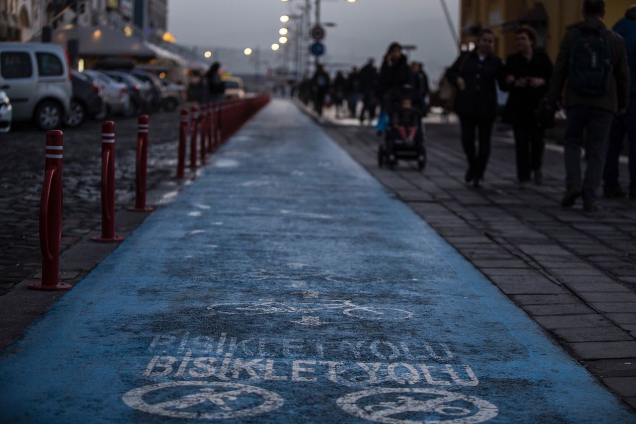 transportation, street, the way forward, incidental people, focus on foreground, city, diminishing perspective, road, large group of people, day, bollard, outdoors, city life, vanishing point, travel, text, mode of transport, guidance, road marking