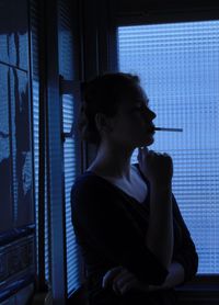 Side view of woman smoking cigarette while resting by window
