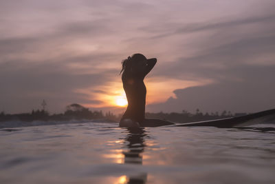 Young woman sitting on surfboard in sea against sky during sunset