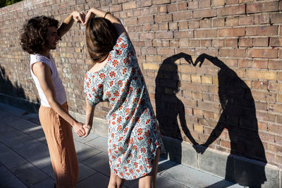 Young couple making heart shape in front of wall on sunny day