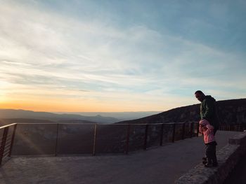 Father with daughter standing on retaining wall against sky during sunset