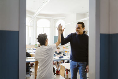 Happy male teacher giving high-five to schoolboy standing in classroom