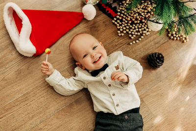 A charming baby is lying on the floor next to a christmas tree with a lollipop 