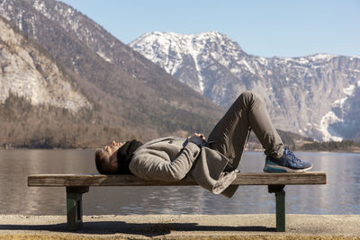 Young man lying outdoors on bench and enjoying mountains, snow, good weather, blue sky, sun