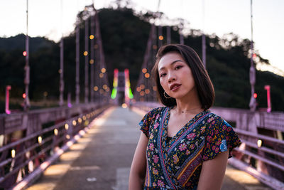 Portrait of beautiful young woman standing against illuminated bridge