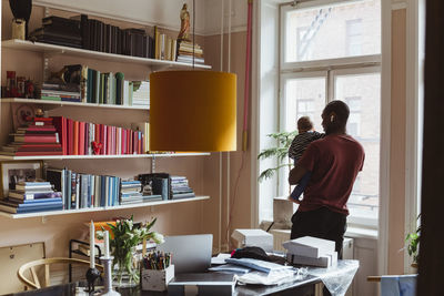 Male entrepreneur carrying male toddler while standing at home office