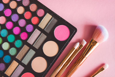 Directly above shot of multi colored eyeshadow palette and brushes on pink background