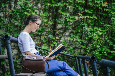 Side view of young woman using phone while sitting on railing