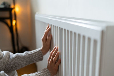 Closeup of woman in sweater puts hands on room central heating battery to warm up in cold apartment