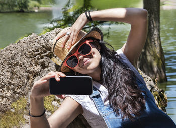 Smiling woman taking selfie with mobile phone against lake during sunny day