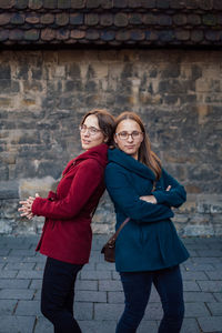 Portrait of smiling sisters standing back to back against stone wall