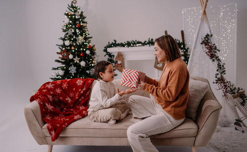 Happy mother sitting on couch at home congratulating her little kid giving surprise gift