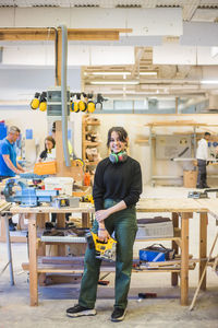 Full length portrait of smiling young female trainee holding power tool while standing in workshop