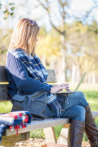 Woman using laptop while sitting on bench against trees