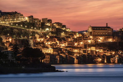 Illuminated buildings by sea against sky at sunset