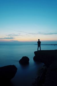 Rear view of silhouette mid adult man standing on cliff by sea against blue sky during sunset