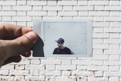 Cropped hand holding photograph against white brick wall during sunny day