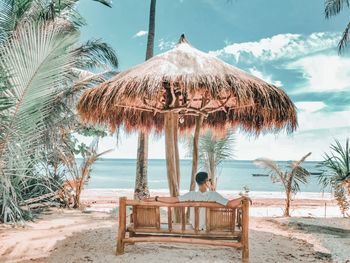 Rear view of young man relaxing on bench at beach against sky