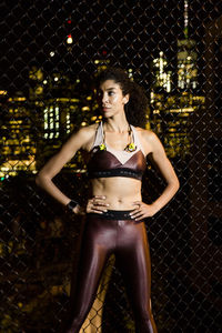 Female athlete with hands on hip standing by fence against illuminated buildings at night