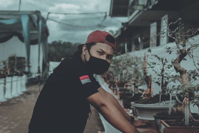 Side view portrait of young man wearing cap and mask standing by potted plant