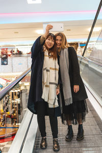 Multi-ethnic beautiful couple of girlfriends on the escalators take a selfie with a smartphone