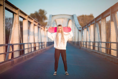 Portrait of young woman holding skateboard while standing on footbridge during sunset