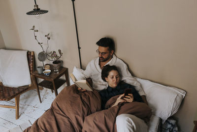 High angle view of man reading book by non-binary person using mobile phone on bed at home