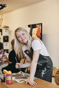 Portrait of smiling female mature student holding paintbrush at table in art class